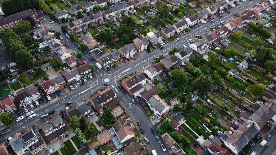 An aerial view of a residential neighbourhood featuring a mix of houses with varied rooftops, some with tiled and others with flat roofs, interspersed with lush green trees and gardens. The streets ar